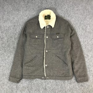 May include: A gray herringbone jacket with a cream-colored, sherpa-lined collar. The jacket has two chest pockets, two lower pockets, and a button-up front. The jacket has a small brand label on the chest.