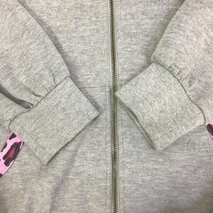 May include: Close-up of a gray zip-up hoodie with ribbed cuffs and a gold zipper. The sleeves are visible, with a pink and black leopard print accent on the left sleeve.