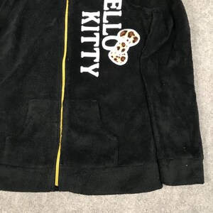 Pu&ograve; includere: Giacca con zip in pile nero con cerniera gialla e logo "Hello Kitty" bianco con fiocco a stampa leopardata.