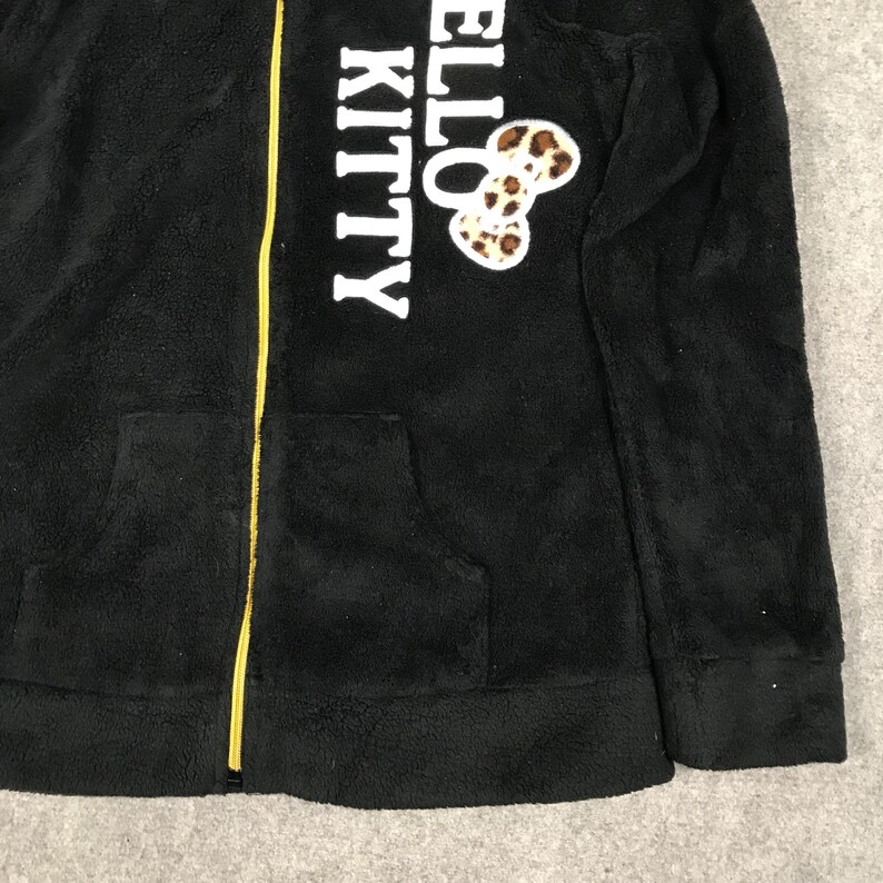 Pu&ograve; includere: Giacca con zip in pile nero con cerniera gialla e logo "Hello Kitty" bianco con fiocco a stampa leopardata.