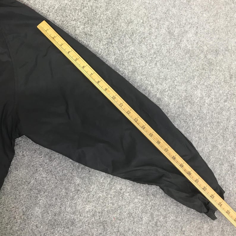 May include: A black long sleeve garment section laid flat next to a yellow wooden ruler. The ruler shows the sleeve length measures approximately 24 inches from the shoulder seam to the cuff edge, providing a clear visual reference for size.