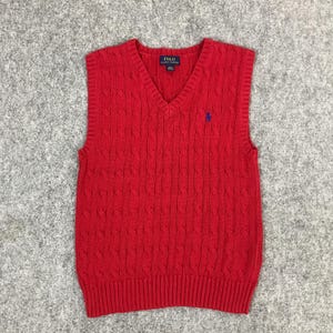 Könnte beinhalten: Rote Strickweste mit Zopfmuster und V-Ausschnitt. Die Weste hat ein kleines blaues Logo auf der Brust und ein Etikett im Nacken. Die Weste ist aus weichem Strickmaterial gefertigt.