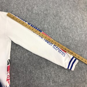 以下が含まれることがあります： 袖に青と赤のアクセントが入った白い長袖シャツ。袖には青色の「THE MARSH ELEMENTS」の文字と、袖口には青と白のストライプが入っています。木製の定規がシャツの上に置かれています。