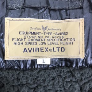 Może przedstawiać: Zbliżenie beżowej etykiety z odzieży lotniczej Avirex. Etykieta zawiera czarny tekst, w tym "EQUIPMENT-TYPE-AVIREX" i "HIGH SPEED LOW LEVEL FLIGHT". Odzież jest ciemnoszara z teksturowaną podszewką.