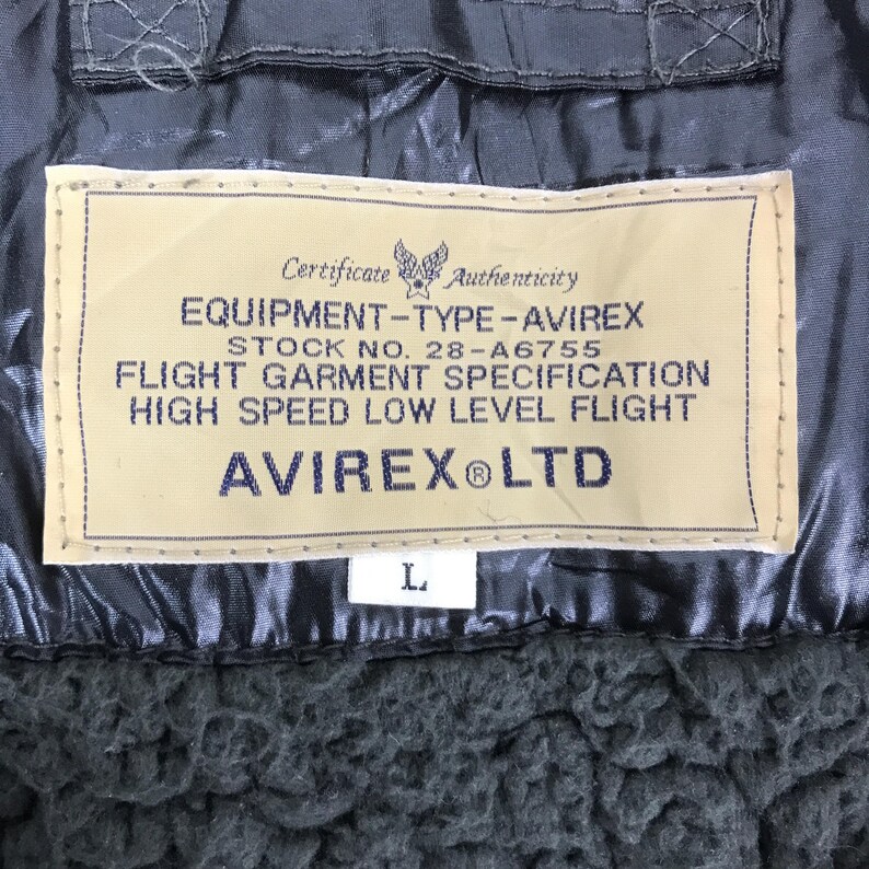 Może przedstawiać: Zbliżenie beżowej etykiety z odzieży lotniczej Avirex. Etykieta zawiera czarny tekst, w tym "EQUIPMENT-TYPE-AVIREX" i "HIGH SPEED LOW LEVEL FLIGHT". Odzież jest ciemnoszara z teksturowaną podszewką.