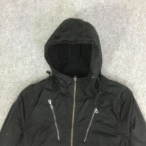 May include: A black hooded jacket featuring a central full-length metal zipper and two diagonal zippered pockets on the chest. The garment includes a drawstring hood with metal eyelets and a smooth, lightweight fabric construction throughout.