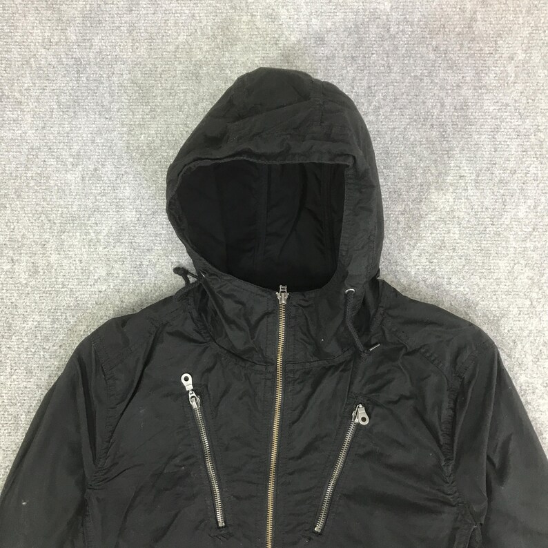 May include: A black hooded jacket featuring a central full-length metal zipper and two diagonal zippered pockets on the chest. The garment includes a drawstring hood with metal eyelets and a smooth, lightweight fabric construction throughout.