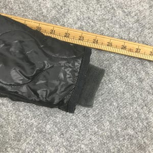 Może przedstawiać: Zbliżenie czarnego pikowanego rękawa kurtki ze ściągaczem. Drewniana linijka jest umieszczona po przekątnej na kurtce, z widocznymi pomiarami od 51 do 71 cm. Kurtka ma błyszczącą, gładką fakturę.