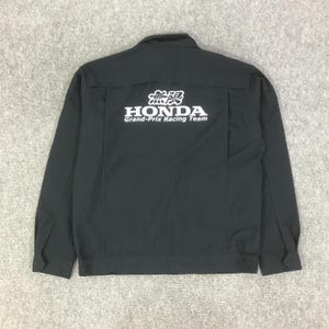 May include: A black long-sleeve jacket featuring a white graphic print on the back that reads "無限 HONDA Grand-Prix Racing Team". The jacket has a collared neckline, long sleeves with cuffs, and a straight hemline with elasticated side panels.