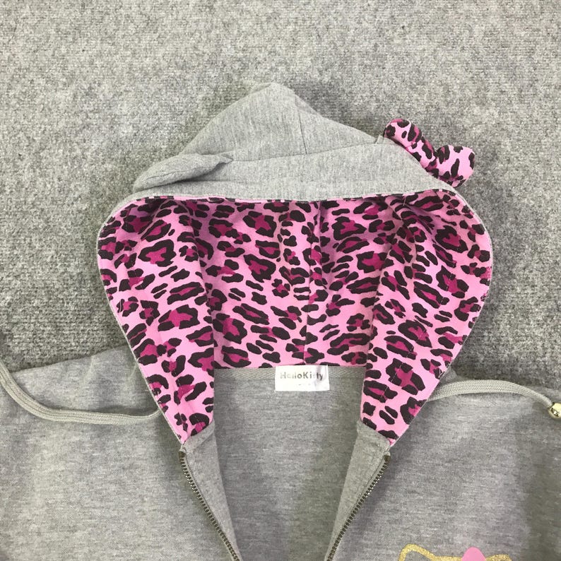 May include: A gray zip-up hoodie with a hood featuring a pink and black leopard print lining. The hood has small, rounded ears and a bow detail. The hoodie has a full zipper and a small tag that says "Hello Kitty".