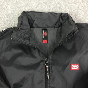 May include: A black zip-up jacket featuring a high collar and a front zipper closure. The interior neck label displays the text Ecko Function and the size L. A red rectangular patch with a white rhinoceros logo is on the chest area.