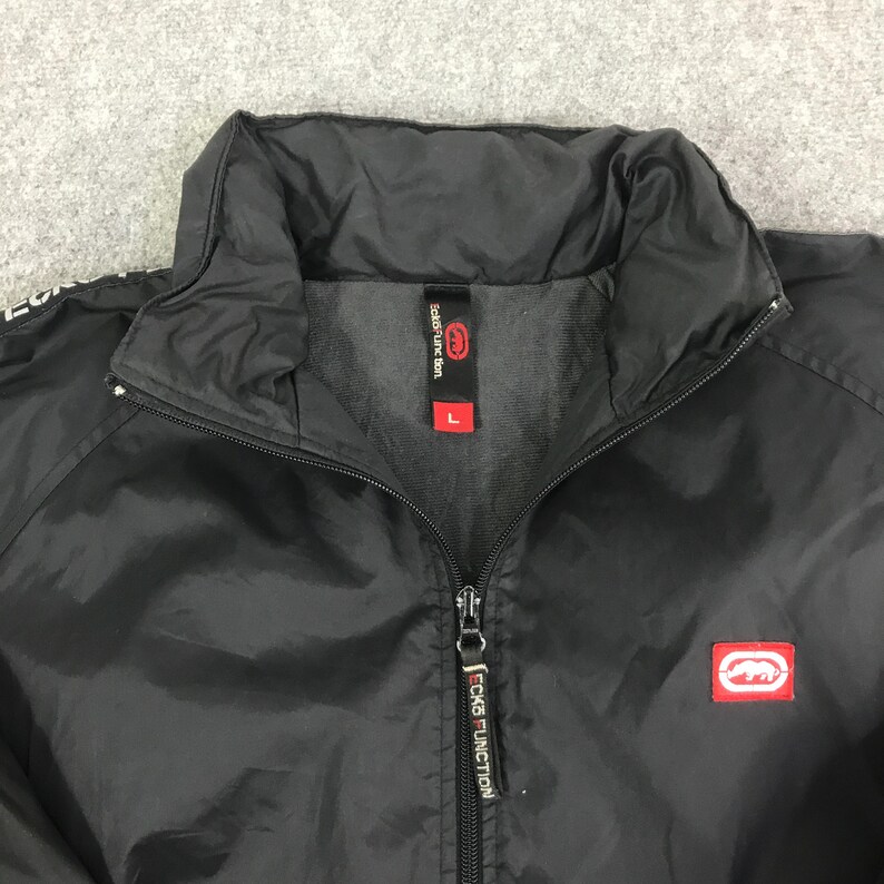 May include: A black zip-up jacket featuring a high collar and a front zipper closure. The interior neck label displays the text Ecko Function and the size L. A red rectangular patch with a white rhinoceros logo is on the chest area.