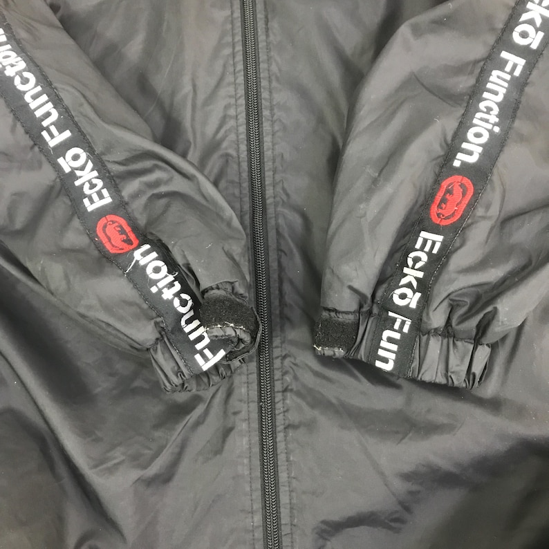 May include: A black zip-up jacket featuring long sleeves with black and white branded tape down the arms. The tape displays the text Ecko Function in white lettering alongside a red circular logo. The cuffs include adjustable black velcro straps.