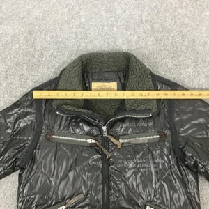 Może przedstawiać: Czarna pikowana kurtka z kołnierzem z ciemnozielonego sztucznego futra. Kurtka posiada zapięcie na zamek błyskawiczny, wiele kieszeni oraz tekst "AVIREX AIRDALE" i "BOILER MAKER UNION N.Y.C. ANCHOR NAVAL SHIP YARD".