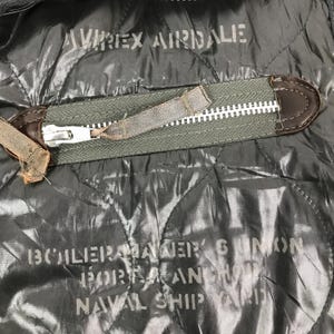 Może przedstawiać: Zbliżenie na ciemnoszarą pikowaną kurtkę z zapięciem na zamek błyskawiczny. Kurtka ma napis "AVIREX AIRDALE" u g&oacute;ry i "BOILERMAKER'S GUNION PORTLAND NAVAL SHIP YARD" na dole. Suwak jest w kolorze oliwkowym.