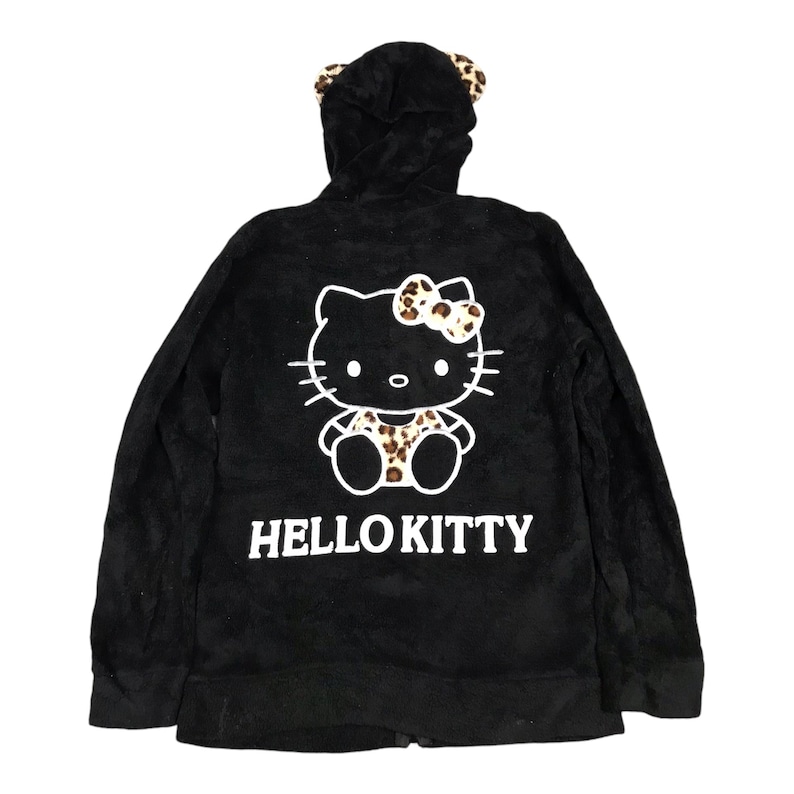 Pu&ograve; includere: Felpa con cappuccio in pile nero con una grafica di Hello Kitty con un fiocco a stampa leopardo e un outfit a stampa leopardo. La felpa ha un cappuccio con un bordo in pelliccia sintetica nera.