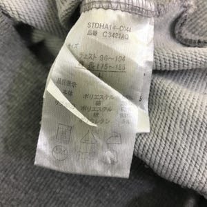 Może przedstawiać: Zbliżenie metki odzieżowej z japońskim tekstem i symbolami pielęgnacji. Etykieta zawiera kod produktu STDHA14-C044 i informacje o rozmiarze. Skład materiału to poliester, bawełna i spandex.