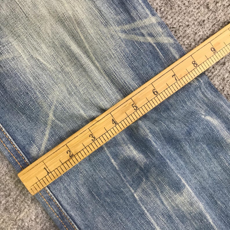 K&ouml;nnte beinhalten: Nahaufnahme einer hellblauen Jeans mit einem Holzlineal, das den Stoff misst. Das Lineal ist in Zentimetern markiert und zeigt Messungen von 2.5 bis 23 cm. Die Jeans hat verblasste Bereiche und sichtbare N&auml;hte.