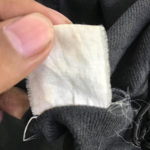 May include: A rectangular white fabric garment care label with frayed edges attached to a dark charcoal gray textile. The label is blank and shows no visible text, branding, or care instructions, featuring a simple woven texture.