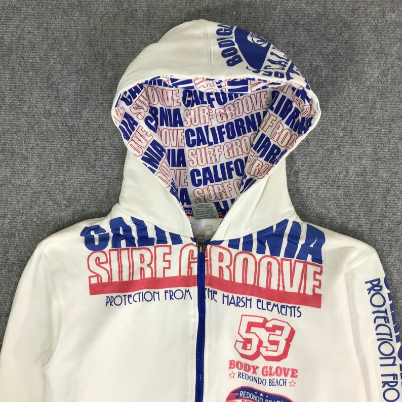 以下が含まれることがあります： 白のジップアップパーカー。青と赤の文字で「CALIFORNIA SURF GROOVE」と「PROTECTION FROM THE HARSH ELEMENTS」と書かれています。フードの裏地には「CALIFORNIA SURF GROOVE」の繰り返し模様があります。「53」の数字が前面にプリントされています。