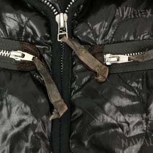 Może przedstawiać: Zbliżenie czarnej pikowanej kurtki ze srebrnymi zamkami i brązowymi sk&oacute;rzanymi detalami. Kurtka ma błyszczącą, odblaskową powierzchnię. Widoczna jest nazwa marki "AVIREX".