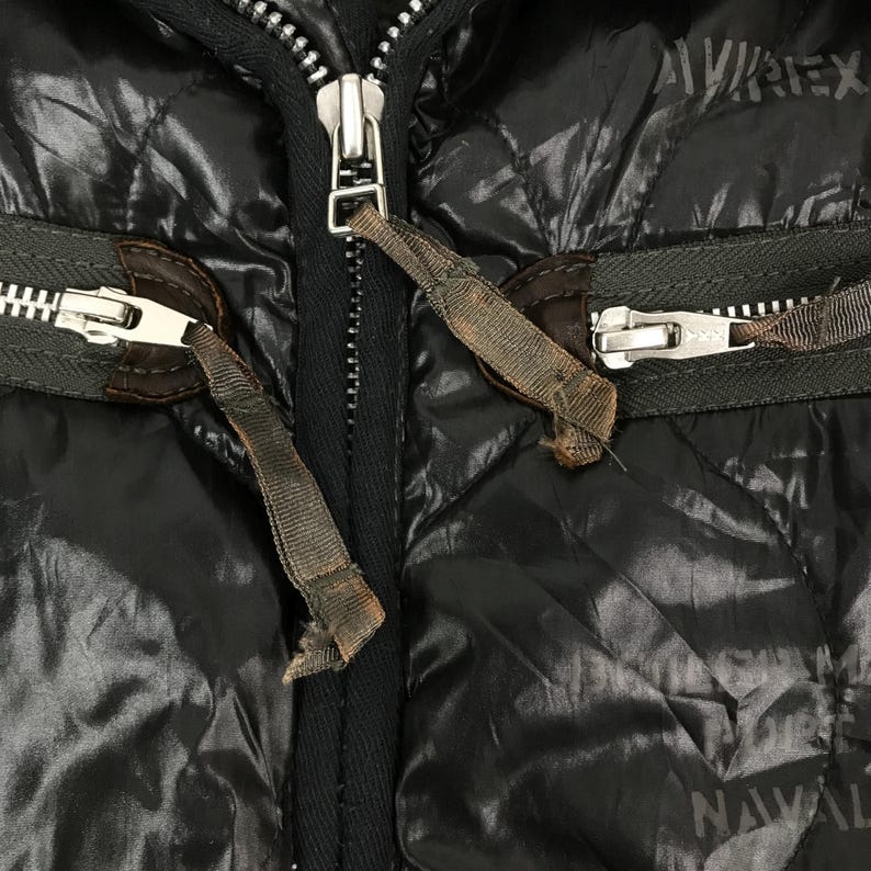 Może przedstawiać: Zbliżenie czarnej pikowanej kurtki ze srebrnymi zamkami i brązowymi sk&oacute;rzanymi detalami. Kurtka ma błyszczącą, odblaskową powierzchnię. Widoczna jest nazwa marki "AVIREX".