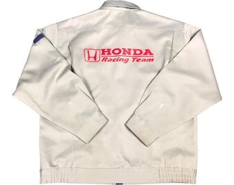 May include: Cream-colored Honda Racing Team jacket with a red logo on the back. The jacket has a zip-up front, long sleeves, and a ribbed hem. A blue accent is visible on the left shoulder.