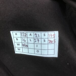 May include: A dark brown fabric surface featuring a white rectangular tag with a grid of handwritten measurements. The grid contains the following text: K 138, A 21, S 17.5, B 16, L 27, S 24.5, W 490, I, W, O. The tag is attached to the dark brown material.