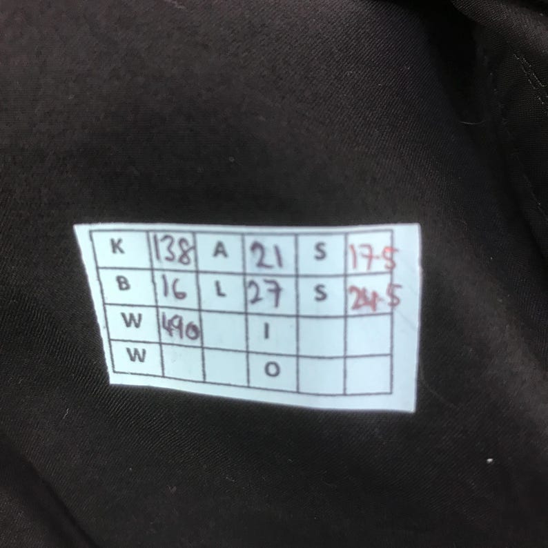 May include: A dark brown fabric surface featuring a white rectangular tag with a grid of handwritten measurements. The grid contains the following text: K 138, A 21, S 17.5, B 16, L 27, S 24.5, W 490, I, W, O. The tag is attached to the dark brown material.