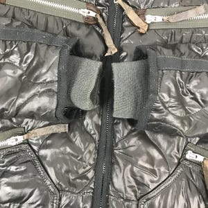 Może przedstawiać: Zbliżenie czarnej pikowanej kurtki z zapięciem na zamek błyskawiczny. Kurtka posiada pikowanie w kształcie romb&oacute;w, prążkowane mankiety i wiele kieszeni na zamek. Zamki mają brązowe sk&oacute;rzane uchwyty. Kurtka wydaje się być wykonana z błyszczącego, wodoodpornego materiału.