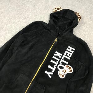 Pu&ograve; includere: Felpa con cappuccio nera in pile con un design Hello Kitty. La felpa ha un cappuccio con orecchie di leopardo pelose e una cerniera dorata. Il testo "Hello Kitty" &egrave; stampato in bianco sul davanti della felpa.