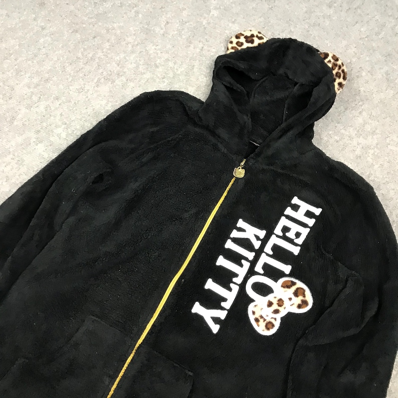 Pu&ograve; includere: Felpa con cappuccio nera in pile con un design Hello Kitty. La felpa ha un cappuccio con orecchie di leopardo pelose e una cerniera dorata. Il testo "Hello Kitty" &egrave; stampato in bianco sul davanti della felpa.