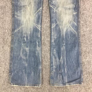 K&ouml;nnte beinhalten: Hellblaue Jeans mit verblichenem, abgenutztem Design. Die Jeans haben einen Bootcut-Stil und zeigen wei&szlig;e, sonnenstrahlenartige Muster. Der Stoff hat ein abgenutztes Aussehen, was auf eine Vintage- oder getragene &Auml;sthetik hindeutet.