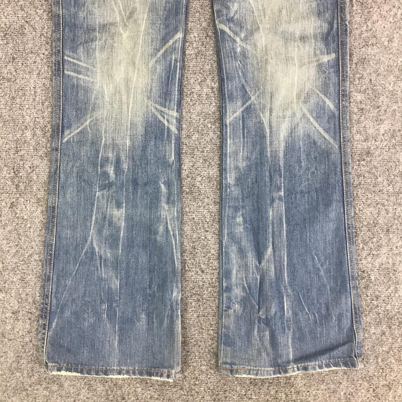 K&ouml;nnte beinhalten: Hellblaue Jeans mit verblichenem, abgenutztem Design. Die Jeans haben einen Bootcut-Stil und zeigen wei&szlig;e, sonnenstrahlenartige Muster. Der Stoff hat ein abgenutztes Aussehen, was auf eine Vintage- oder getragene &Auml;sthetik hindeutet.