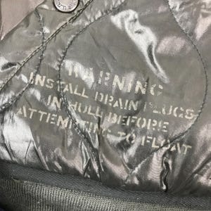 Może przedstawiać: Zbliżenie pikowanej, oliwkowozielonej tkaniny z białym napisem "WARNING". Tekst zawiera instrukcje dotyczące instalacji kork&oacute;w spustowych przed pr&oacute;bą pływania. Tkanina ma błyszczącą jakość.