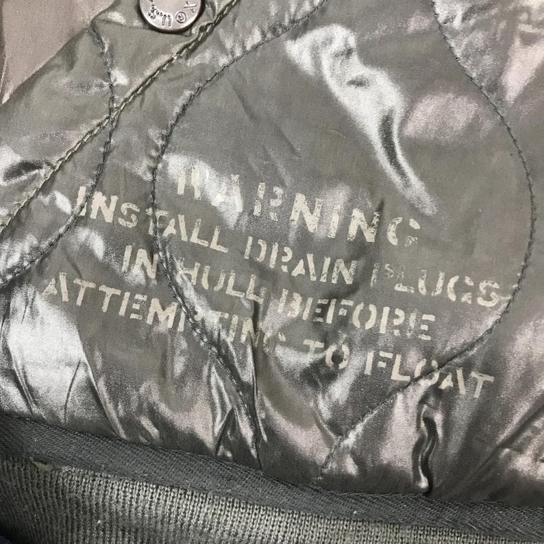 Może przedstawiać: Zbliżenie pikowanej, oliwkowozielonej tkaniny z białym napisem "WARNING". Tekst zawiera instrukcje dotyczące instalacji kork&oacute;w spustowych przed pr&oacute;bą pływania. Tkanina ma błyszczącą jakość.