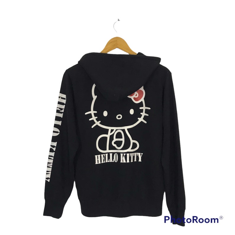 May include: Black hooded sweatshirt with a white Hello Kitty graphic on the back and the words "Hello Kitty" printed below. The left sleeve has the words "Hello Kitty" printed vertically in white.