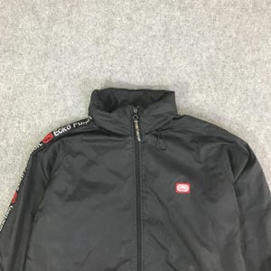 May include: A black zip-up jacket featuring a high collar and a full-length front zipper. The sleeves display a white stripe with the text Ecko Function and a red logo. A small red Ecko Function logo patch is positioned on the left chest area.