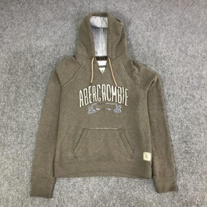 May include: A brown long-sleeve hooded sweatshirt featuring the text ABERCROMBIE in white lettering across the chest. The hoodie includes a front kangaroo pocket, a white and grey striped inner hood lining, and tan drawstring cords at the neck.