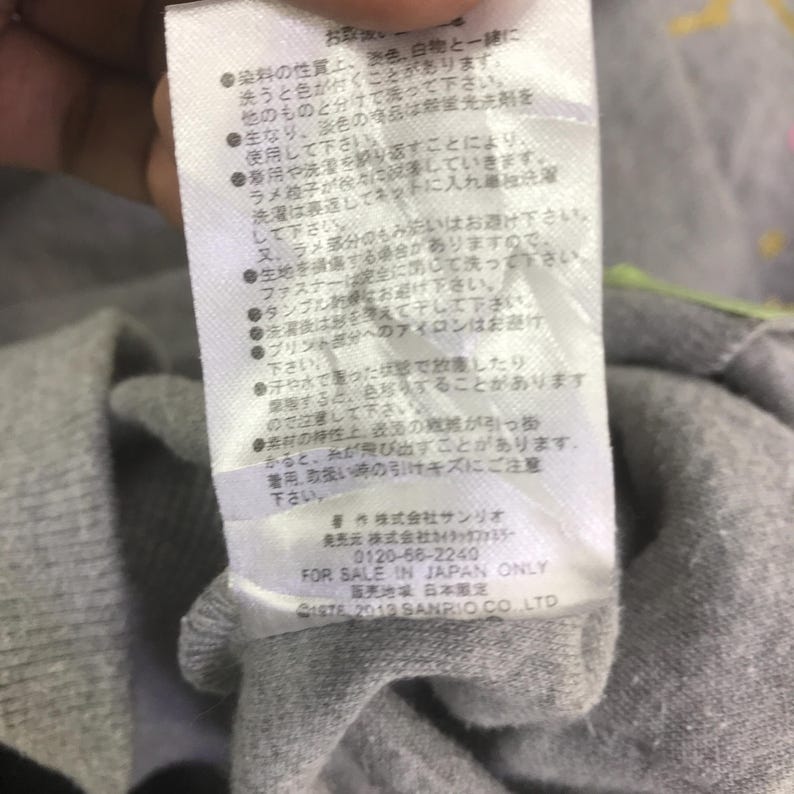 May include: Close-up of a white tag with Japanese text and the text "FOR SALE IN JAPAN ONLY". The tag is attached to a gray fabric. The tag also includes the text "&copy;1976, 2013 SANRIO CO. LTD".