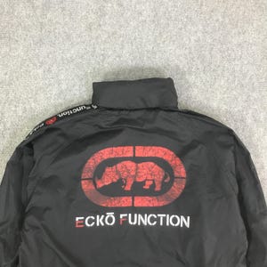 May include: A black windbreaker jacket featuring a high collar and long sleeves. The back displays a large, distressed red rhino logo inside an oval, with the white text ECKO FUNCTION printed below. A black stripe with white text runs along the shoulder.