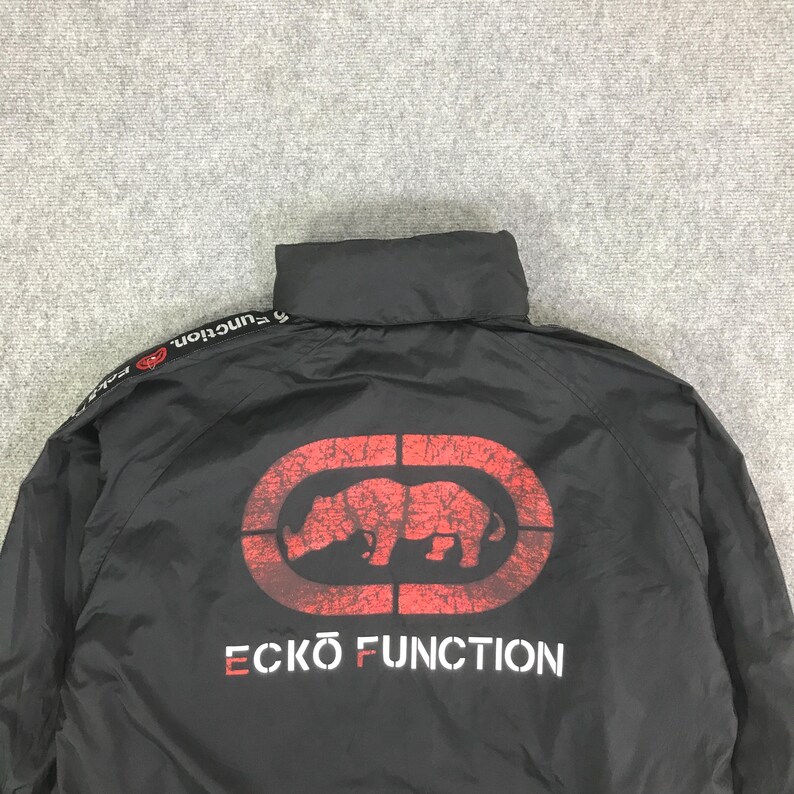 May include: A black windbreaker jacket featuring a high collar and long sleeves. The back displays a large, distressed red rhino logo inside an oval, with the white text ECKO FUNCTION printed below. A black stripe with white text runs along the shoulder.