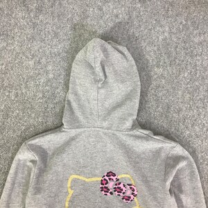 May include: A gray hooded sweatshirt featuring a graphic of a cartoon cat with a pink and black leopard print bow. The hood is a matching gray color. The sweatshirt is made of a soft material.