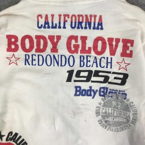 以下が含まれることがあります： 「CALIFORNIA BODY GLOVE REDONDO BEACH 1953」の文字が入った白いTシャツ。テキストは赤、青、黒で表示されています。また、星のグラフィックと「CALIFORNIA SURFSTYLE」の文字が入った円形のグラフィックも特徴です。