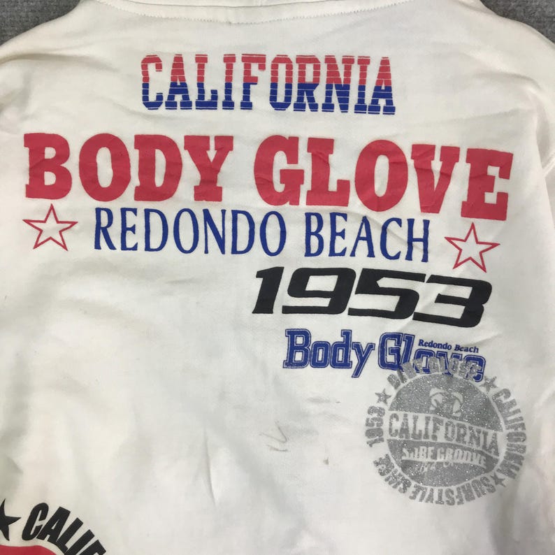 以下が含まれることがあります： 「CALIFORNIA BODY GLOVE REDONDO BEACH 1953」の文字が入った白いTシャツ。テキストは赤、青、黒で表示されています。また、星のグラフィックと「CALIFORNIA SURFSTYLE」の文字が入った円形のグラフィックも特徴です。