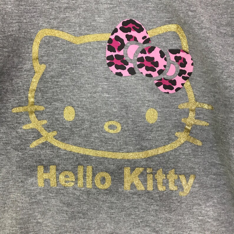 May include: A gray sweatshirt featuring a gold glitter outline of Hello Kitty's face. The bow is pink with black leopard print spots. The words "Hello Kitty" are printed in gold glitter below the face.