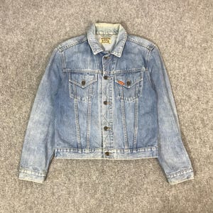 May include: A light blue denim jacket with a classic design. The jacket has a button-up front, two chest pockets, and two side pockets. The collar and cuffs show slight wear. A small red tag is visible on the right chest pocket.