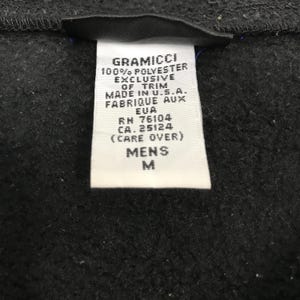 以下が含まれることがあります： Gramicciの衣類のタグのクローズアップ。白いタグには、"100% POLYESTER"と"MADE IN U.S.A."を含む黒い文字が書かれています。また、"MENS"とサイズ"M"も示しています。背景は濃い灰色の生地です。