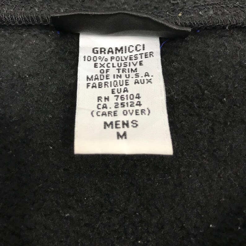 以下が含まれることがあります： Gramicciの衣類のタグのクローズアップ。白いタグには、"100% POLYESTER"と"MADE IN U.S.A."を含む黒い文字が書かれています。また、"MENS"とサイズ"M"も示しています。背景は濃い灰色の生地です。