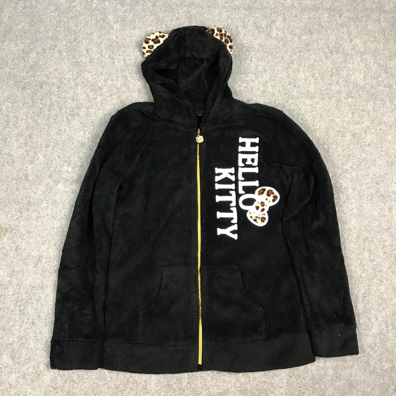 Pu&ograve; includere: Felpa con zip nera in pile con un design Hello Kitty. La felpa ha una grafica Hello Kitty bianca con un fiocco a pois leopardati e orecchie da gatto sul cappuccio.